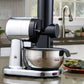 A countertop kitchen scene showing a black and silver electric juicer with a stainless steel bowl, with a plate of food in the foreground and a kitchen sink and window in the background.