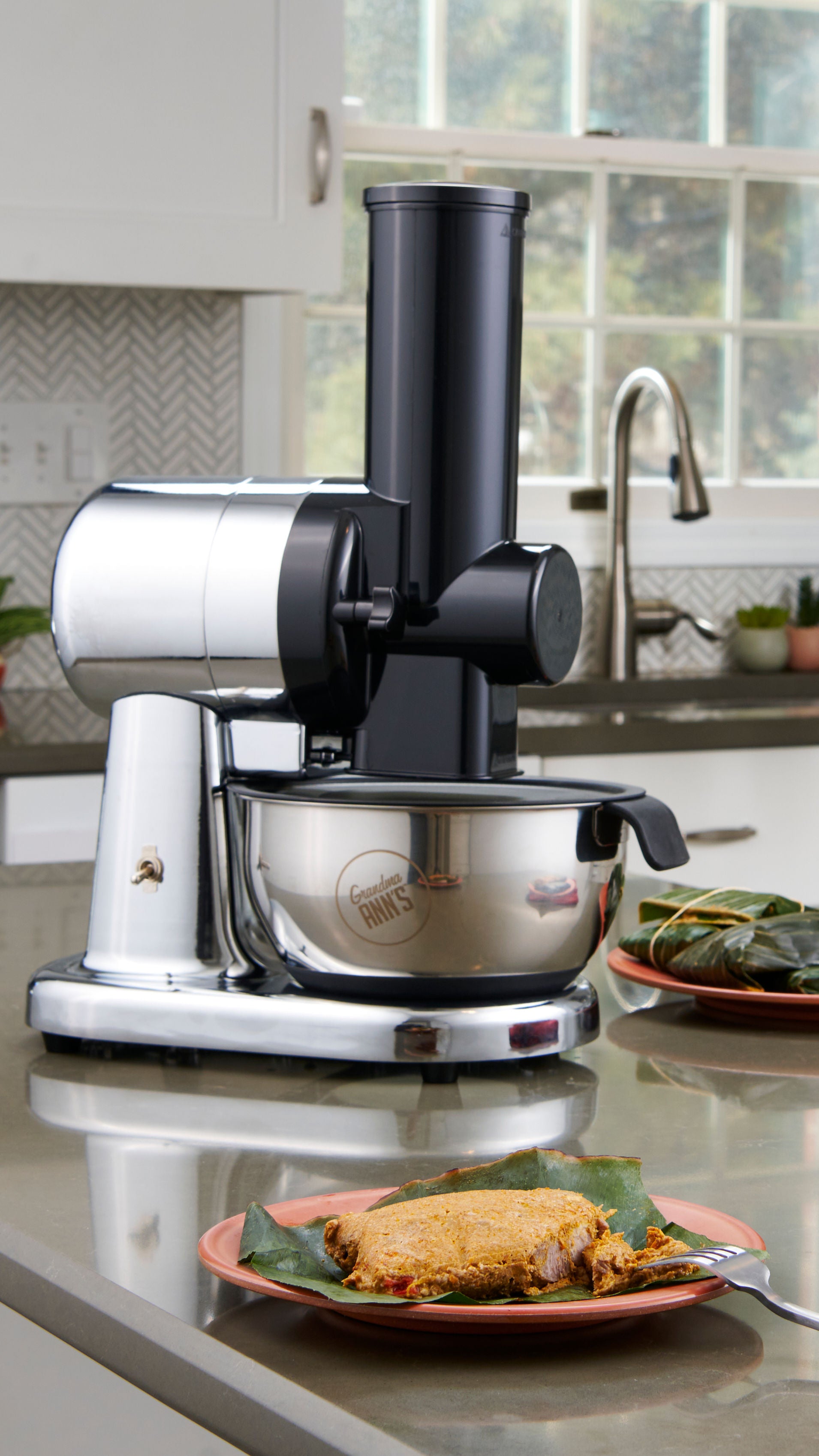 A countertop kitchen scene showing a black and silver electric juicer with a stainless steel bowl, with a plate of food in the foreground and a kitchen sink and window in the background.